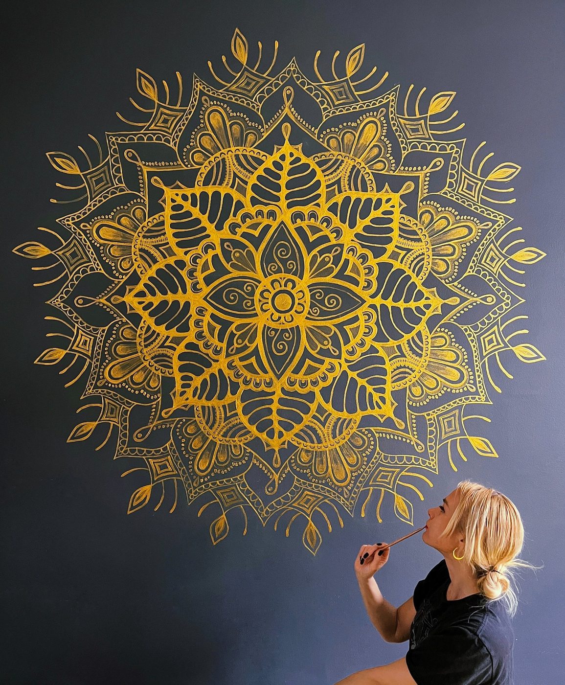 Lasma painting a large gold mandala on a dark wall